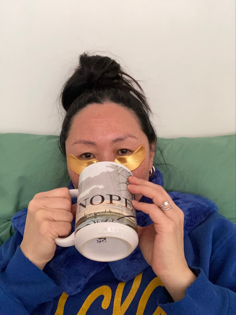 Image of the writer sitting with a heating pad around her neck, gold colored under eye pads, dark hair up in a relaxed bun, wearing her blue college sweatshirt and holding a large mug with the word, "Nope" on it, full of tea. White background. Green pillows.