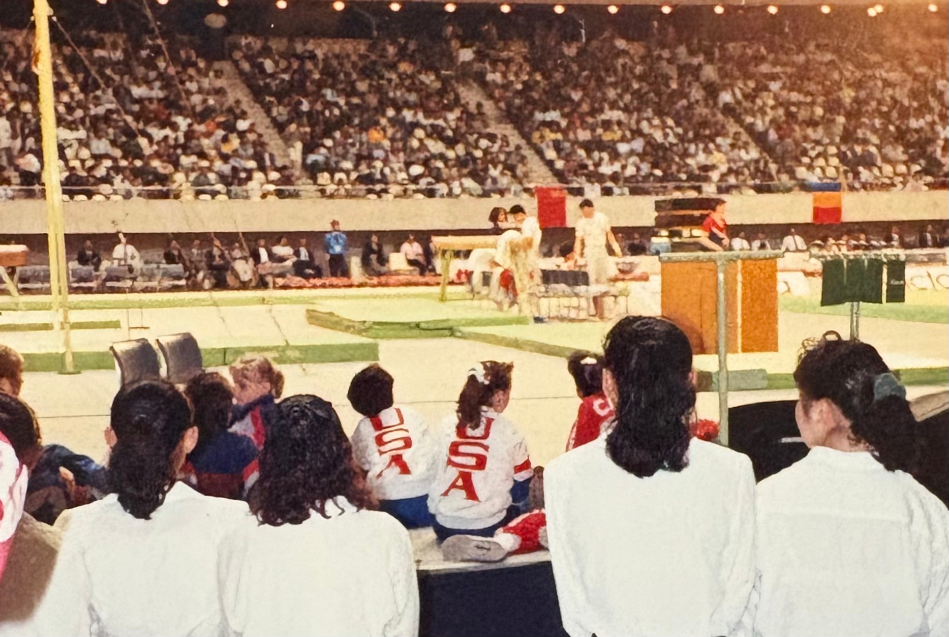 Photo of a gymnastics competition. In the foreground, the backs of three onlookers, each with dark shoulder length hair, wearing white blouses. In front of them, backs of two young USA gymnasts, sitting on the floor, watching the events, wearing their track suits with "USA" in red on the back. In the distance, the gymnastics arena - green floor and beam. In the far distance, the other side of the arena - the stands filled with watchers.