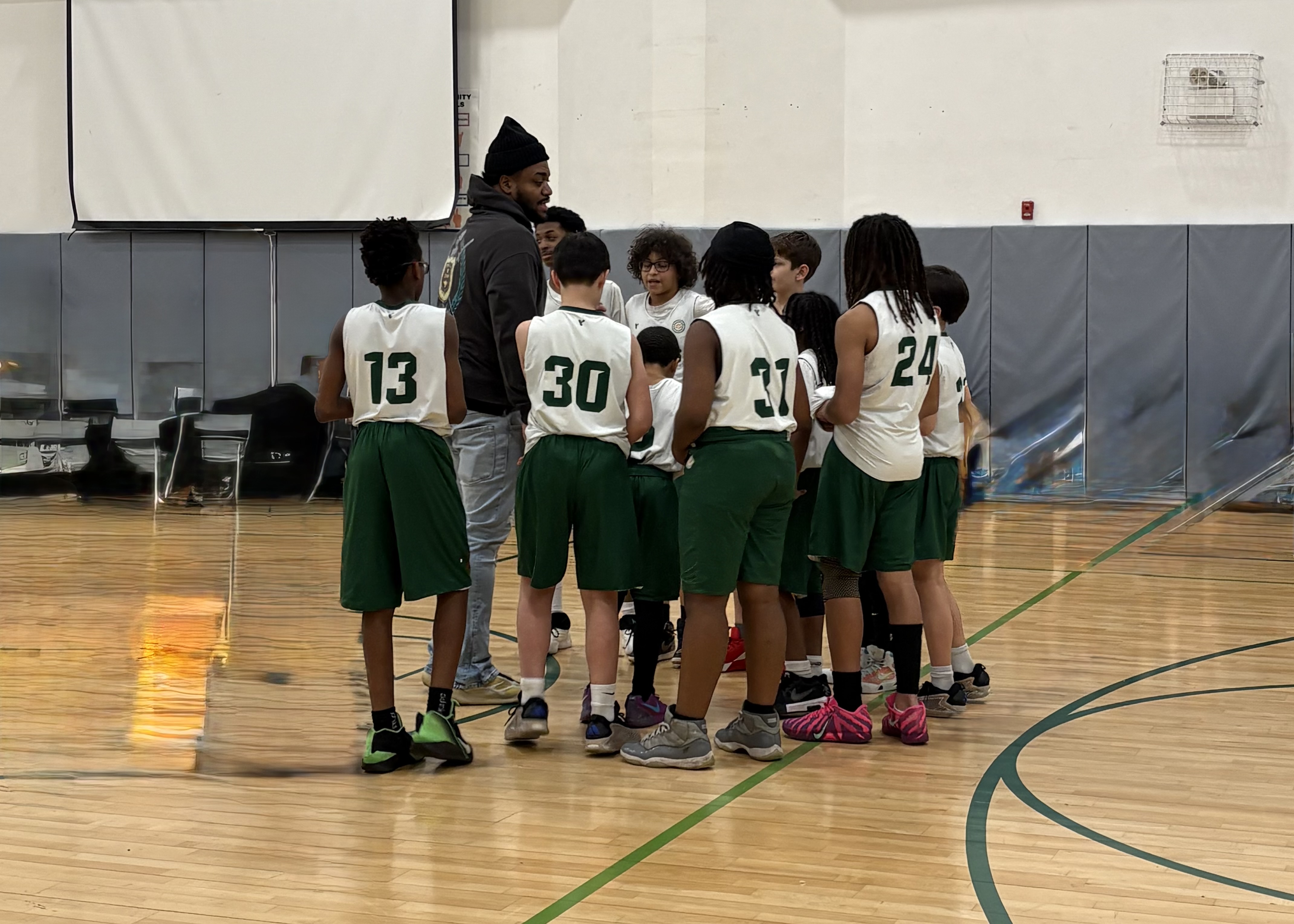 Photo of a coach with the team. Coach is a tall man wearing a black beanie, black hoodie, and jeans, standing with the team surrounding. The team wears dark green shorts with white jerseys with green numbers.
